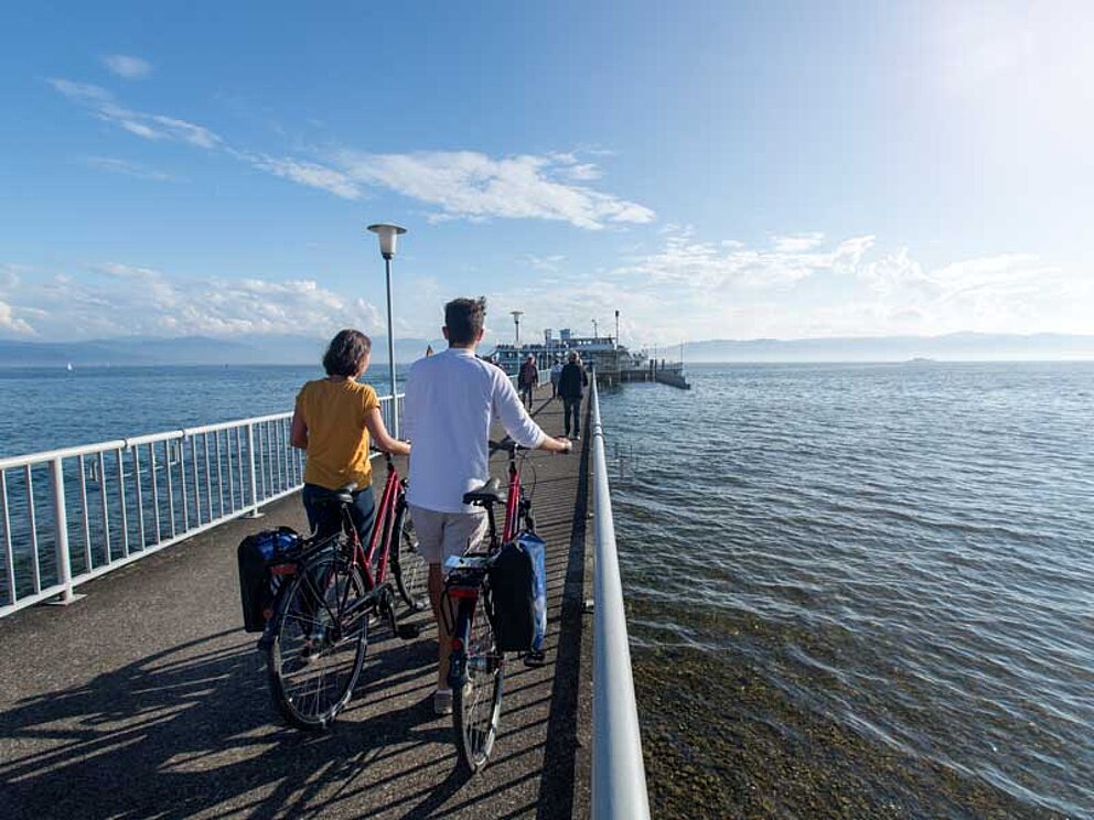 Zwei Radfahrer stehen auf einem Steg und blicken auf den Bodensee