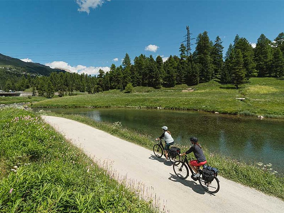 2 Radfahrer am Schotterweg fahren an einem Teich vorbei, im Hintergrund Bäume