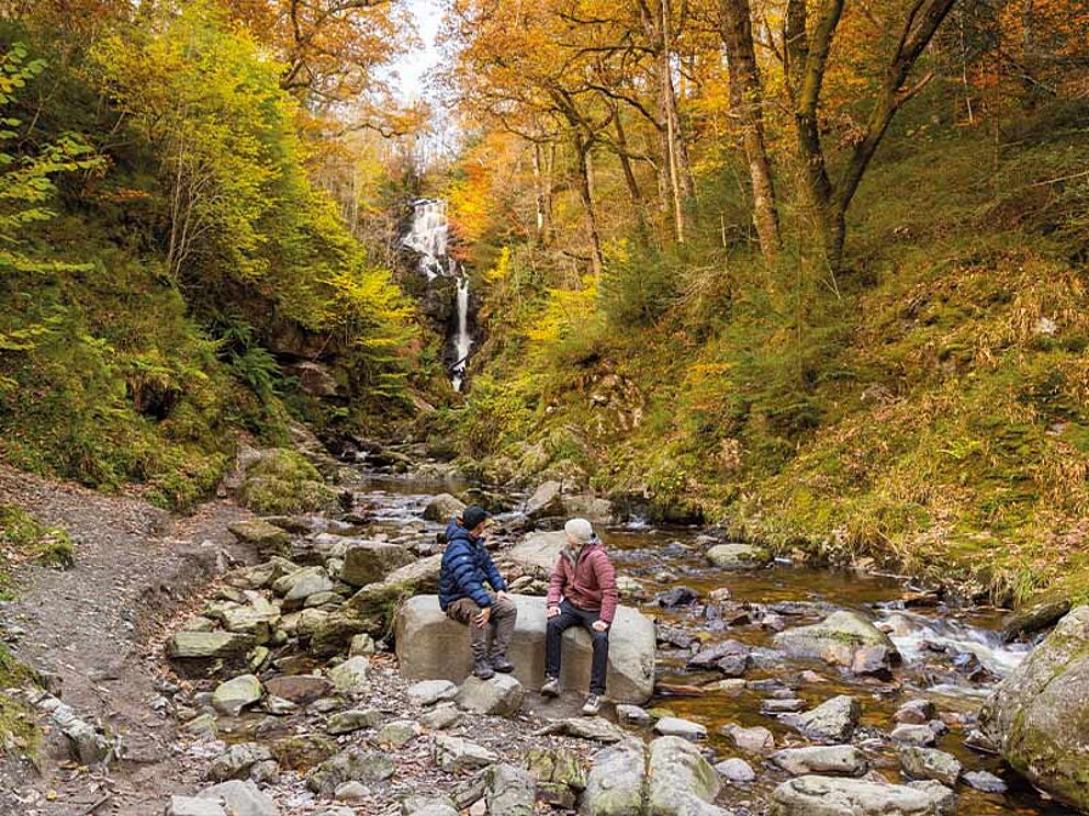 2 Personen sitzen auf großen Steinen vor einem Wasserfall im herbstlichen Queen Elizabeth Park.