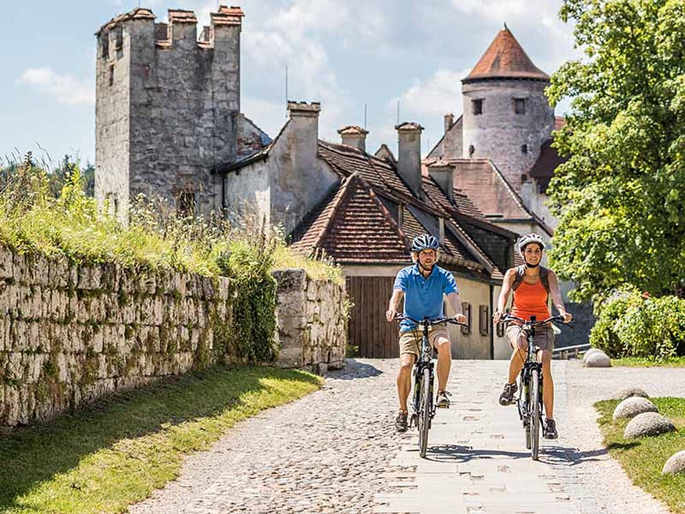 2 Radfahrer auf einem Schotterweg, im Hintergrund eine Burg in Burghausen