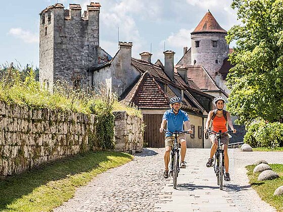 2 Radfahrer auf einem Schotterweg, im Hintergrund eine Burg in Burghausen
