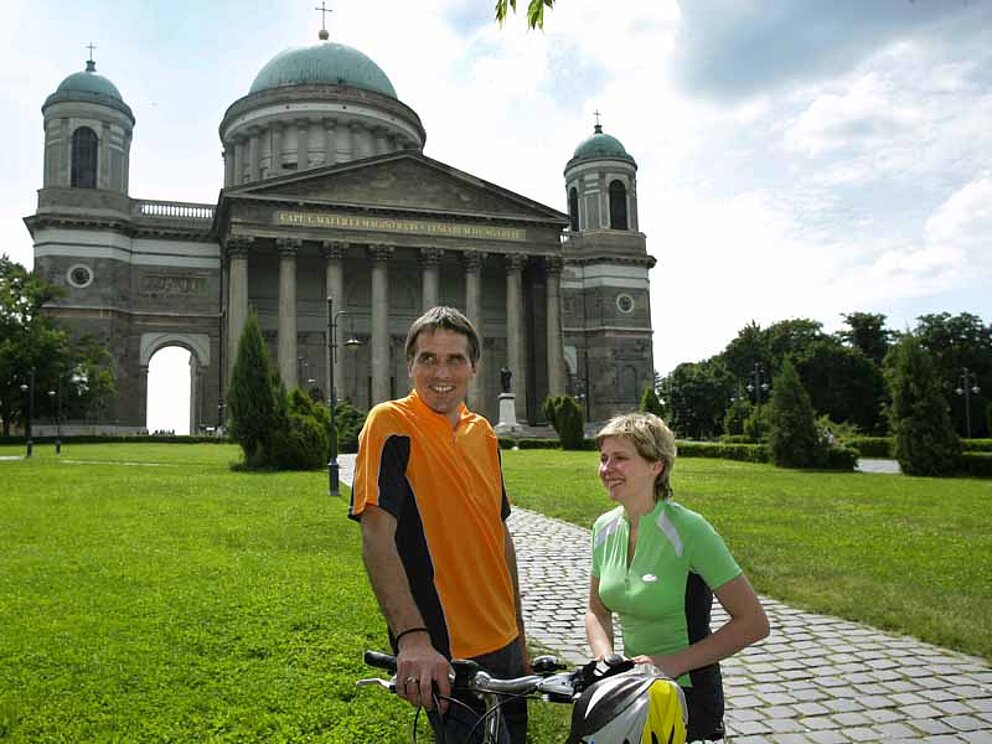 Ein Mann und eine Frau in Radressen stehen vor der Basilika in Esztergom. Das Gebäude hat eine große Kuppel. Links und rechts je ein Türmchen. Die Fassade gleicht einem Portal zu einem griechischen Tempel.