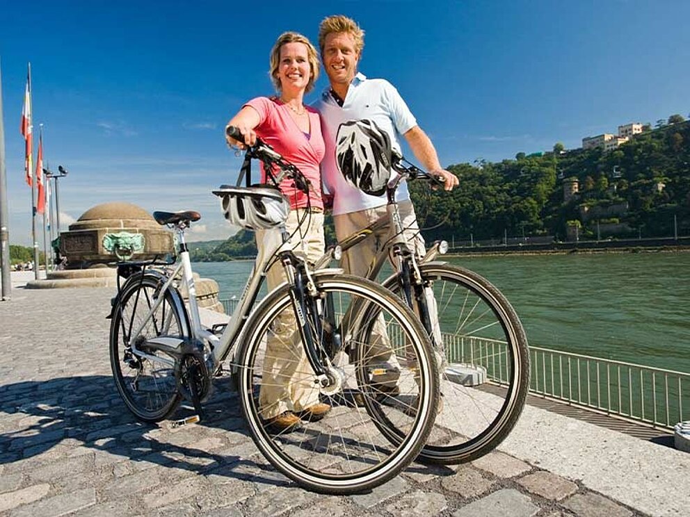 Ein Paar steht bei ihren Fahrrädern, den Helm am Lenker befestigt. Im Hintergrund die Mosel.
