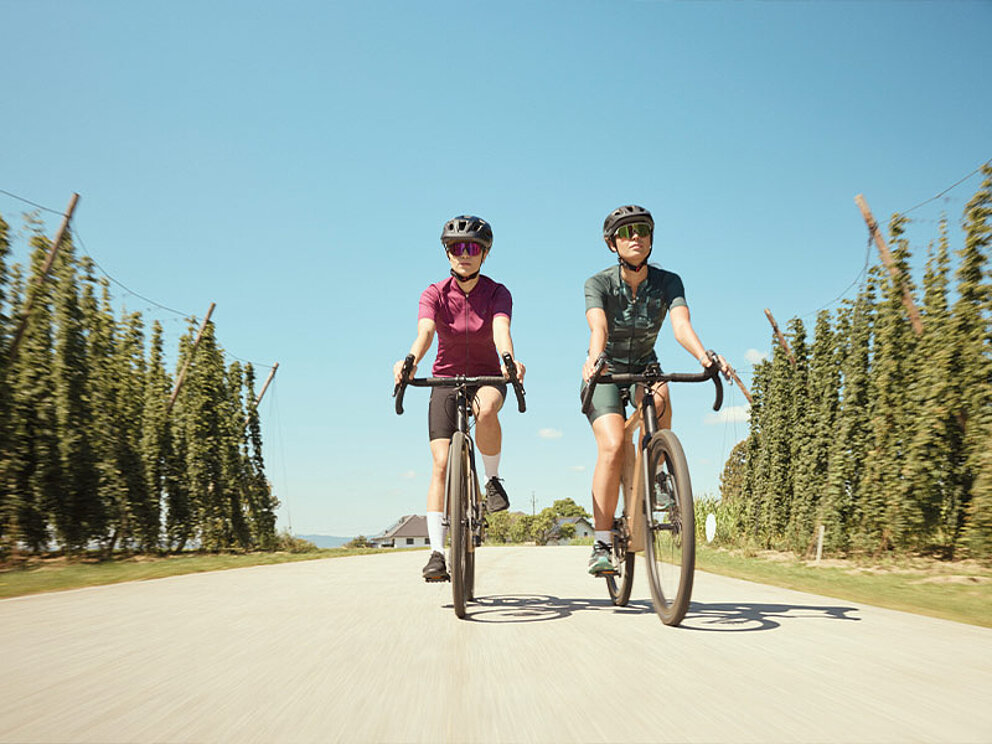 2 Damen auf Gravelbikes fahren auf einem Weg durch Hopfenfelder.