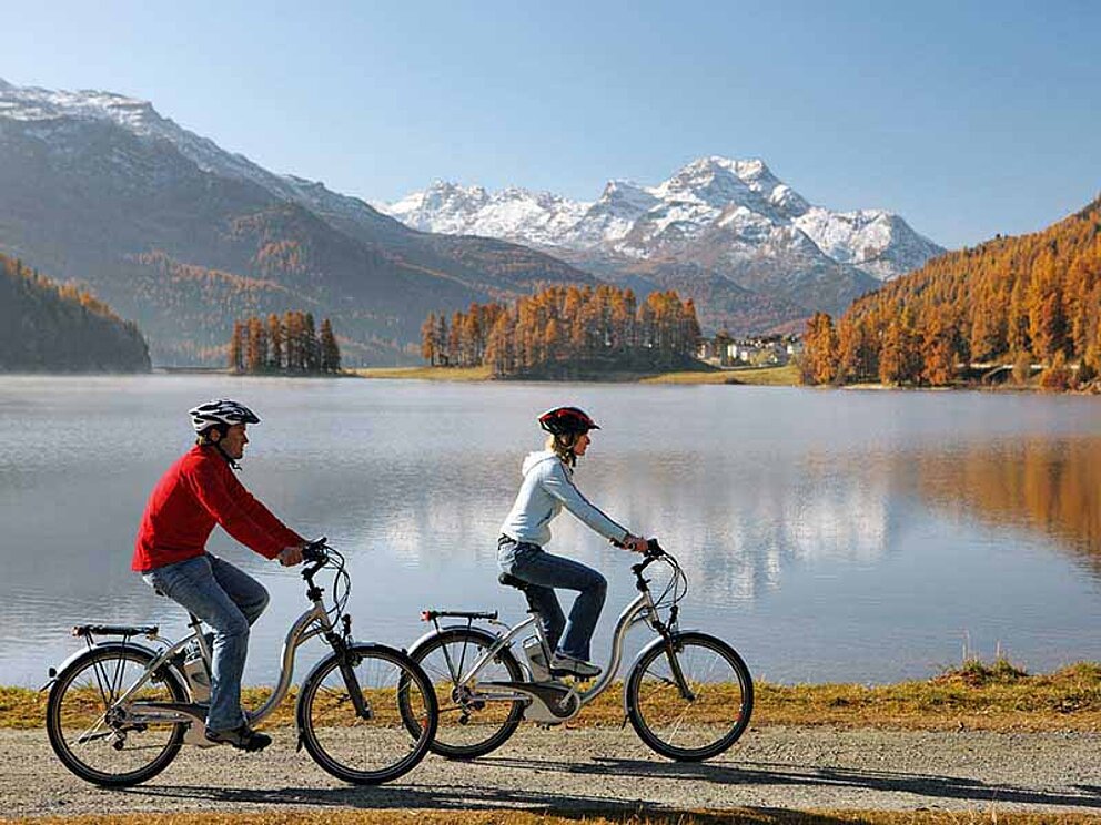 Zwei Radfahrer radeln am Ufer des Silvaplanersee. Im Hintergrund schneebedeckte Alpengipfel.