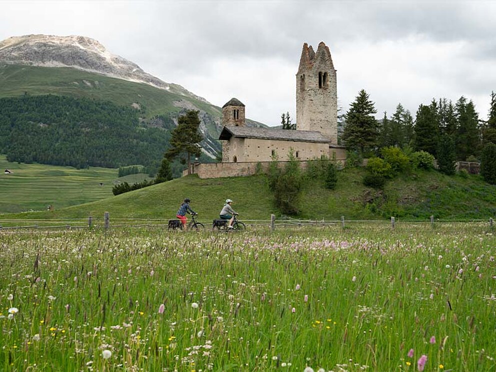 2 Radfahrer, im Vordergrund eine Blumenwiese, im Hintergrund ein Schloss