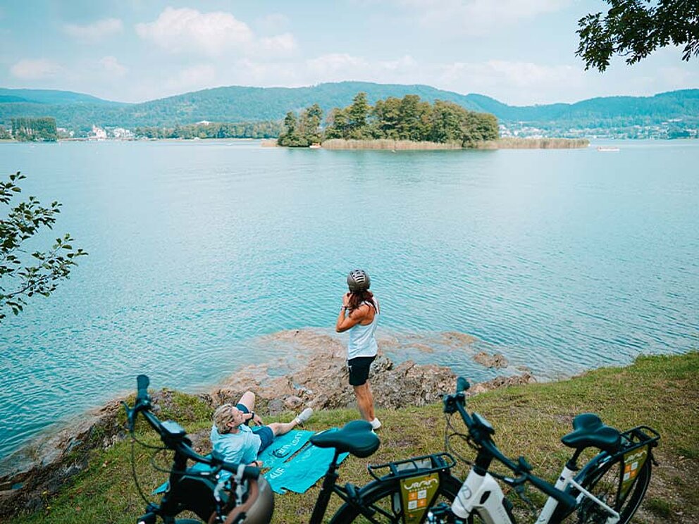 2 Personen am Ufer des Wörthersees, im Vordergrund ihre E-Bikes, im Hintergrund eine Insel im See