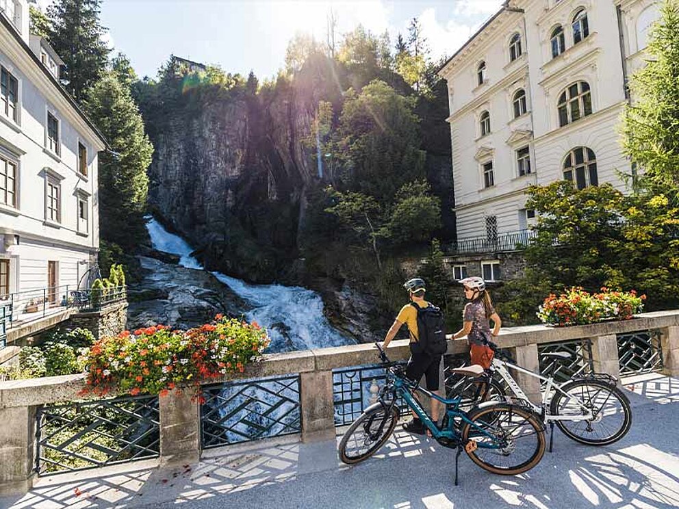 2 Personen mit Fahrrad stehen am Geländer beim Wasserfall im Zentrum von Bad Gastein