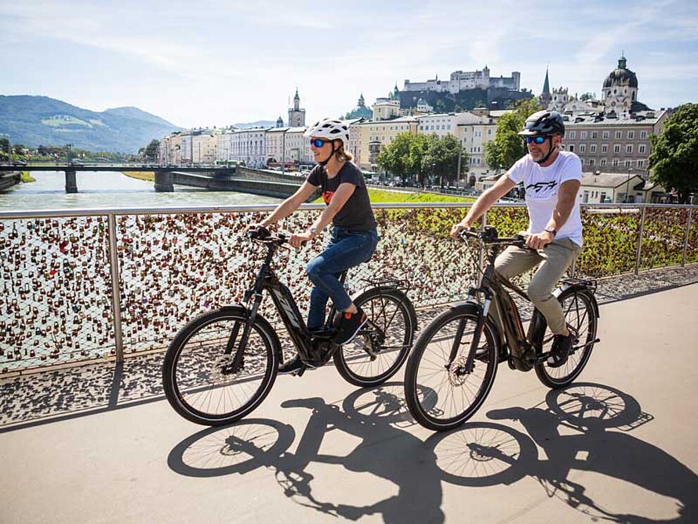 Mann und Frau mit E-Bikes radeln auf der Salzachbrücke in Salzburg, dahinter die Festung Hohensalzburg
