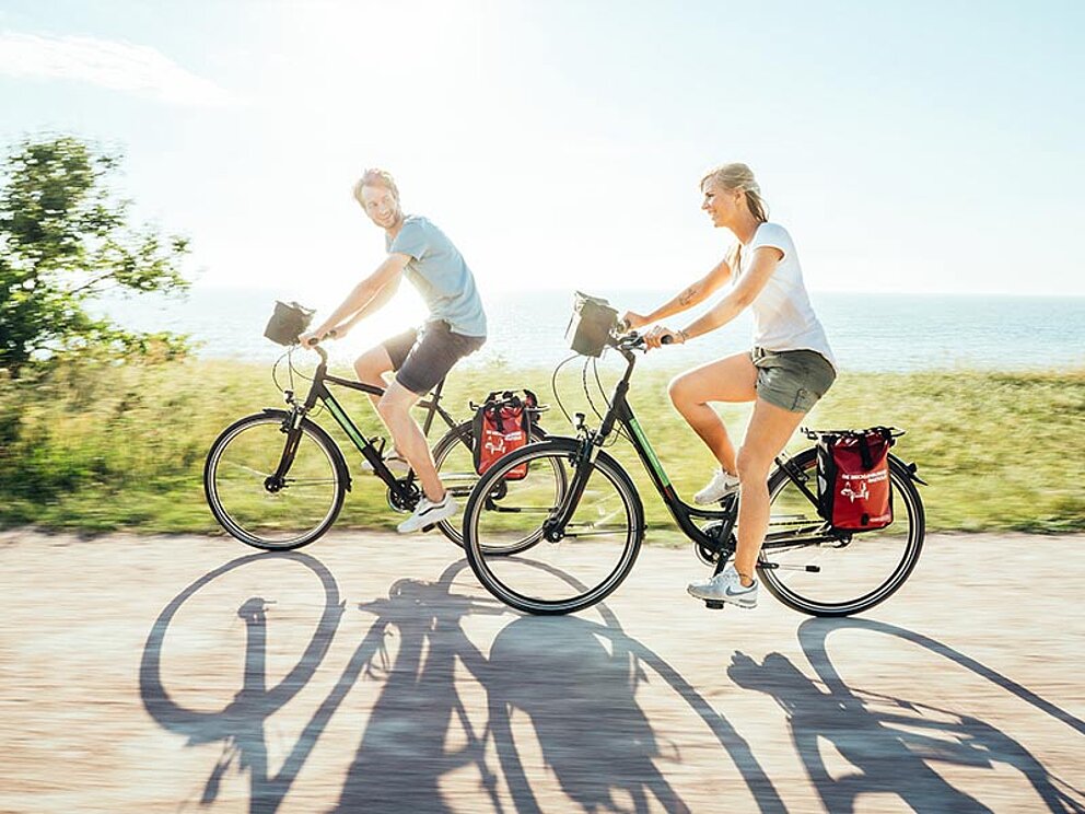 2 Radfahrer am asphaltierten Radweg, im Hintergrune die Ostsee