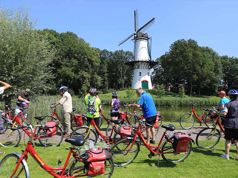 Radfahrer stoppen vor einer weißen Windmühle. Zu sehen sind viele rote Fahrräder im Vordergrund