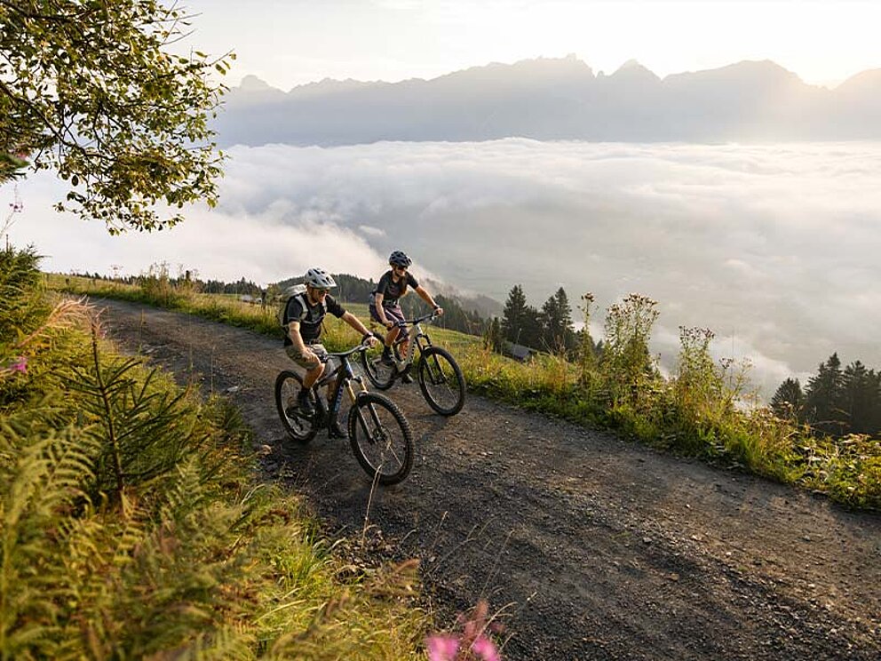 2 Biker auf einem Schotterweg bei Sonnenschein über der Nebelgrenze, im Hintergrund Bergpanorama und die Nebelschicht