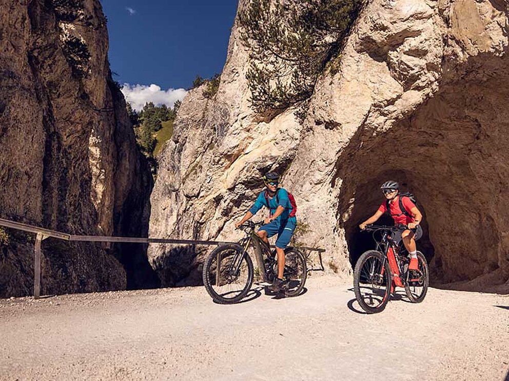 2 Mountainbiker verlassen einen grobgehauenen Gebirgstunnel