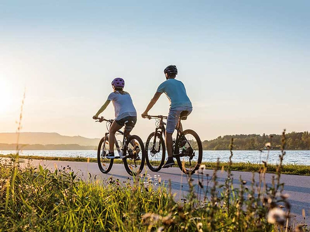 Mann und Frau radeln in den Sonnenuntergang. Die Donau fließt ruhig daneben.