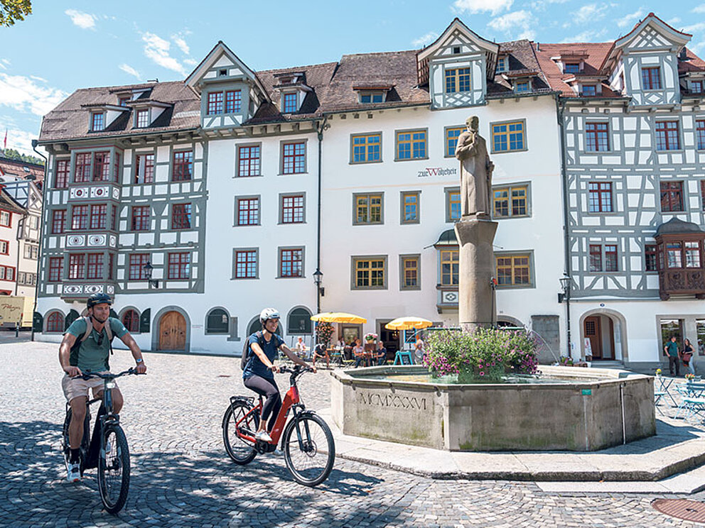2 Radfahrer fahren über einen geplfasterten Marktplatz, vorbei am Brunnen