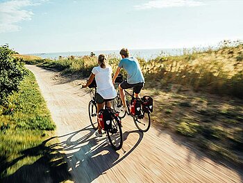 2 Radfahrer am sandigen Radweg, umgeben von Wiese und Dünen