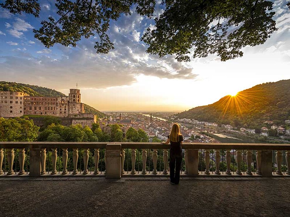 Die Sonne geht über Heidelberg unter. Im Vordergrund eine Dame von hinten, Der Blick schweift über das Schloss und die Altstadt.