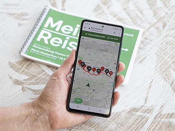 Ein Smartphone zeigt das Radurlaub-Navi, gehalten von einer Person. Im Hintergrund liegt das Routenbuch.