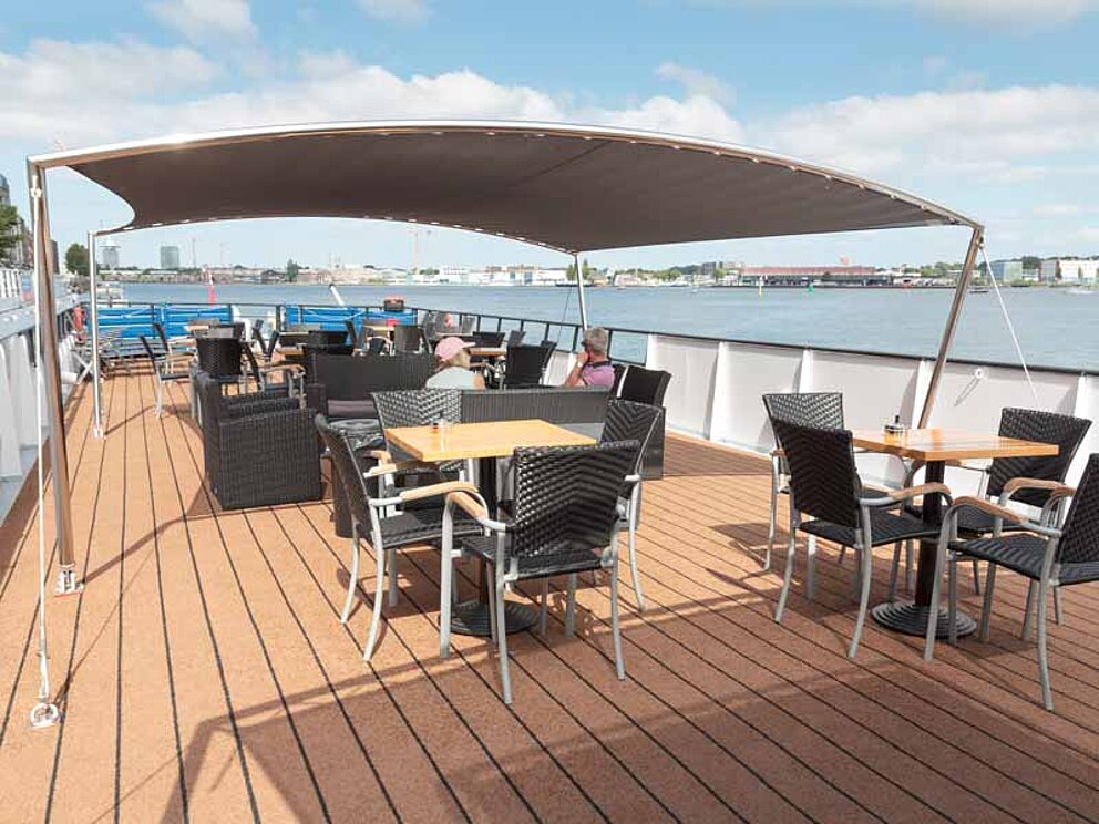 Menschen plaudern an Tischen am Sonnendeck auf einem Schiff. Ein Sonnensegel schützt vor der Sonne