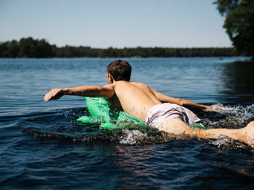 Ein Bub auf einer Krokodil-Luftmatratze plantscht in einem schwedischen See