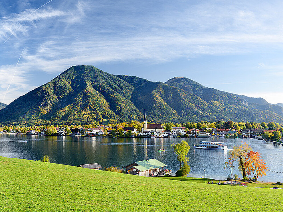 Blick über den Tegernsee auf den Ort Bad Wiessee. Im Vordergrund saftige Wiesen, im Hintergrund ein bewaldeter Berg