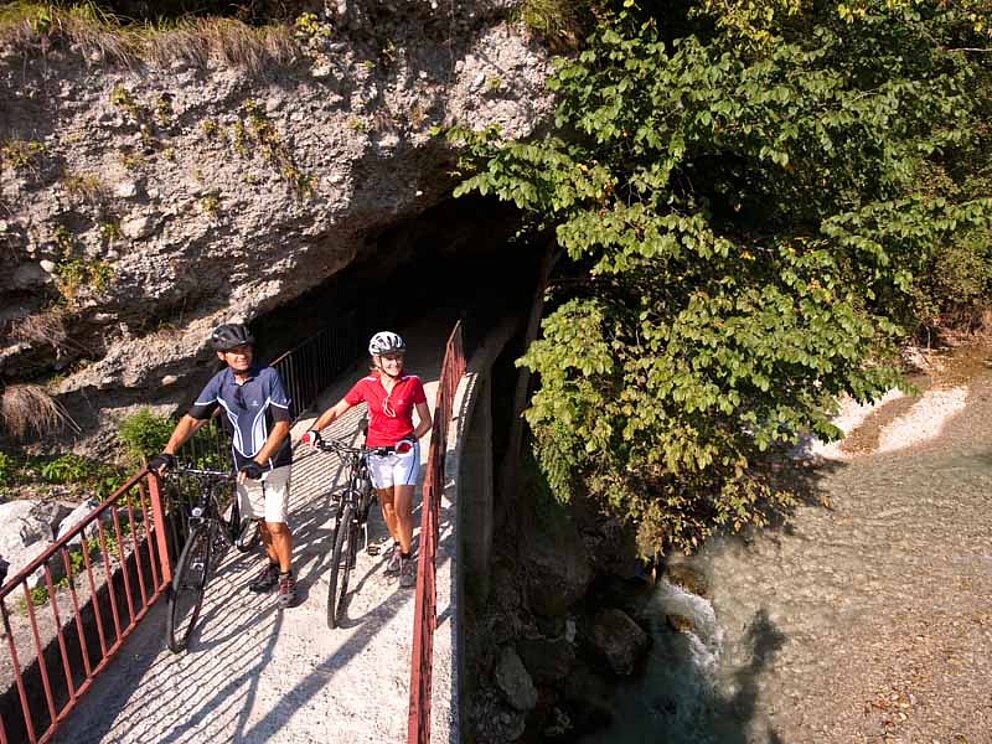 zwei Radfahrer schieben Fahrräder über eine schmale Brücke. Unter Brücke fließt ein Fluss. Die Brücke mündet in einen Tunnel.