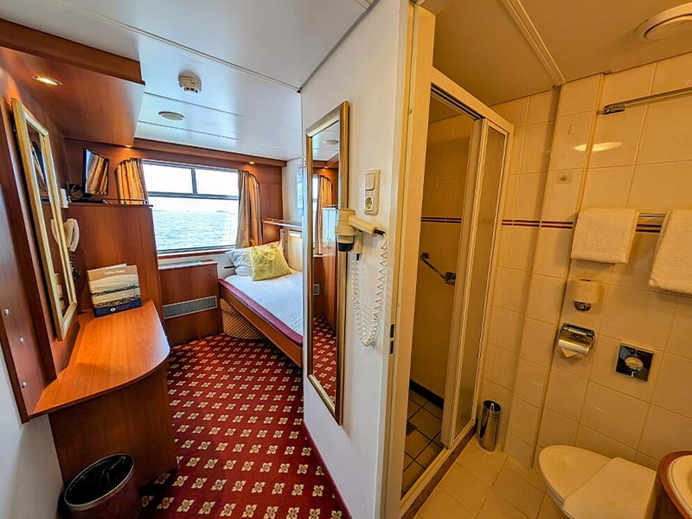Ein Bett, Fenster, Teppichboden, Schreibtisch mit Spiegel, Badnische mit Duschkabine