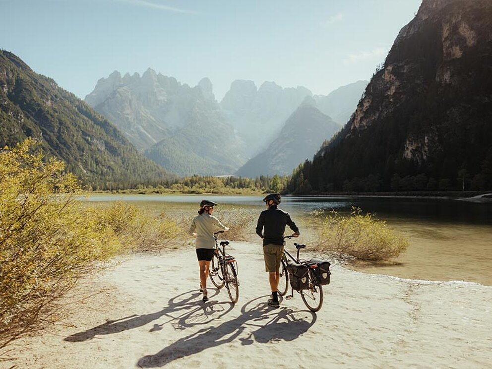 2 Radfahrer von Hinten machen Halt am Ufer eines Bergsees, im Hintergrund Berge