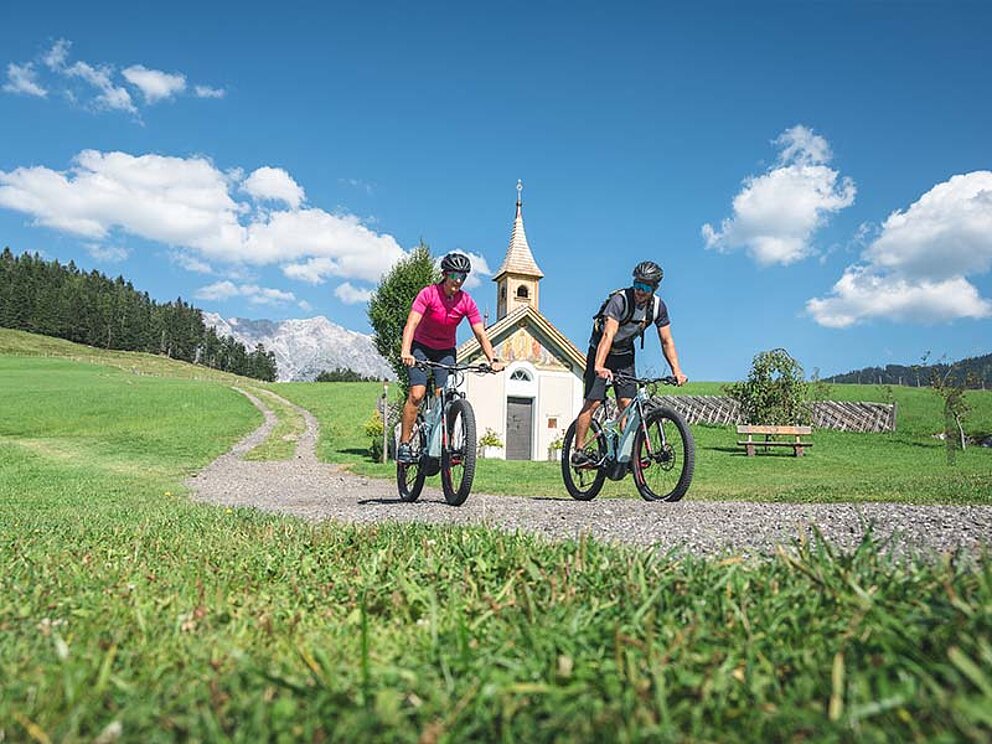 2 Biker auf E-Mountainbikes, im Hintergrund ein kleine Kapelle