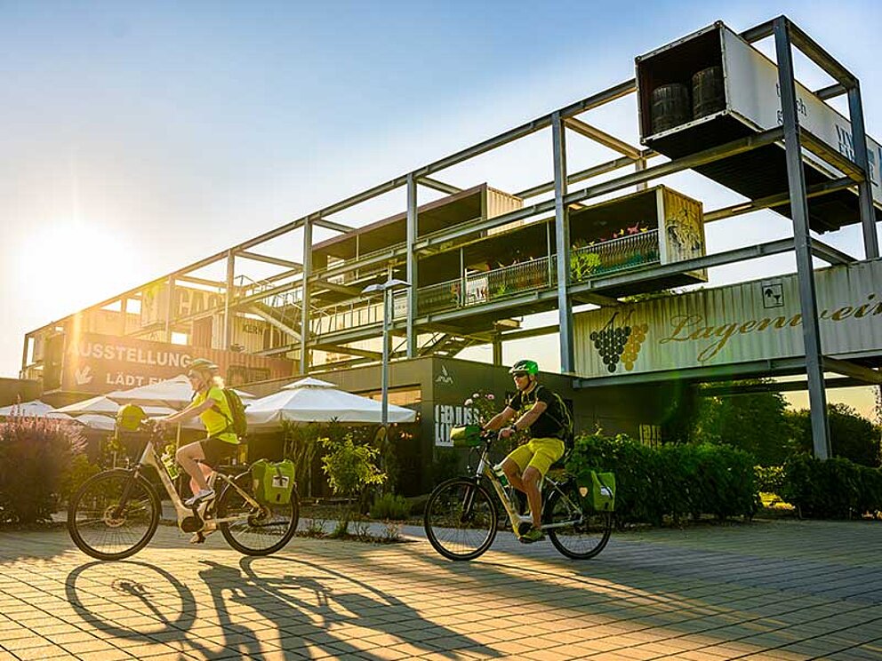 2 Radfahrer radeln am künstlerisch gestalteten Gebäude der Vinofaktur vorbei.