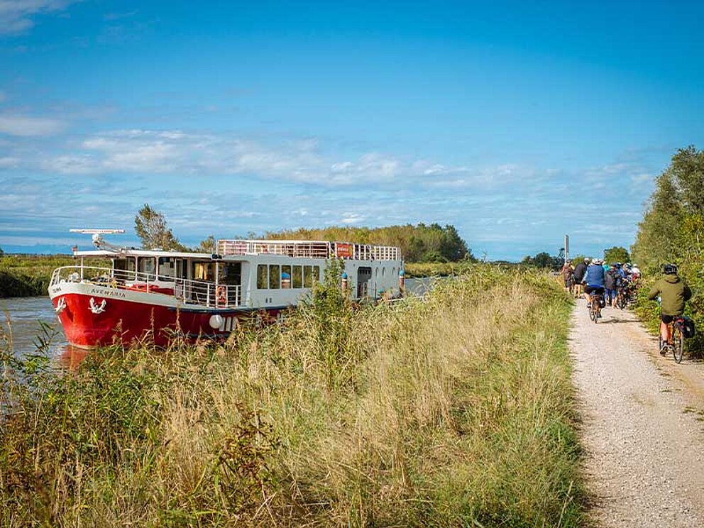 links das Schiff am Fluss, rechts die Radfahrer am Schotterradweg