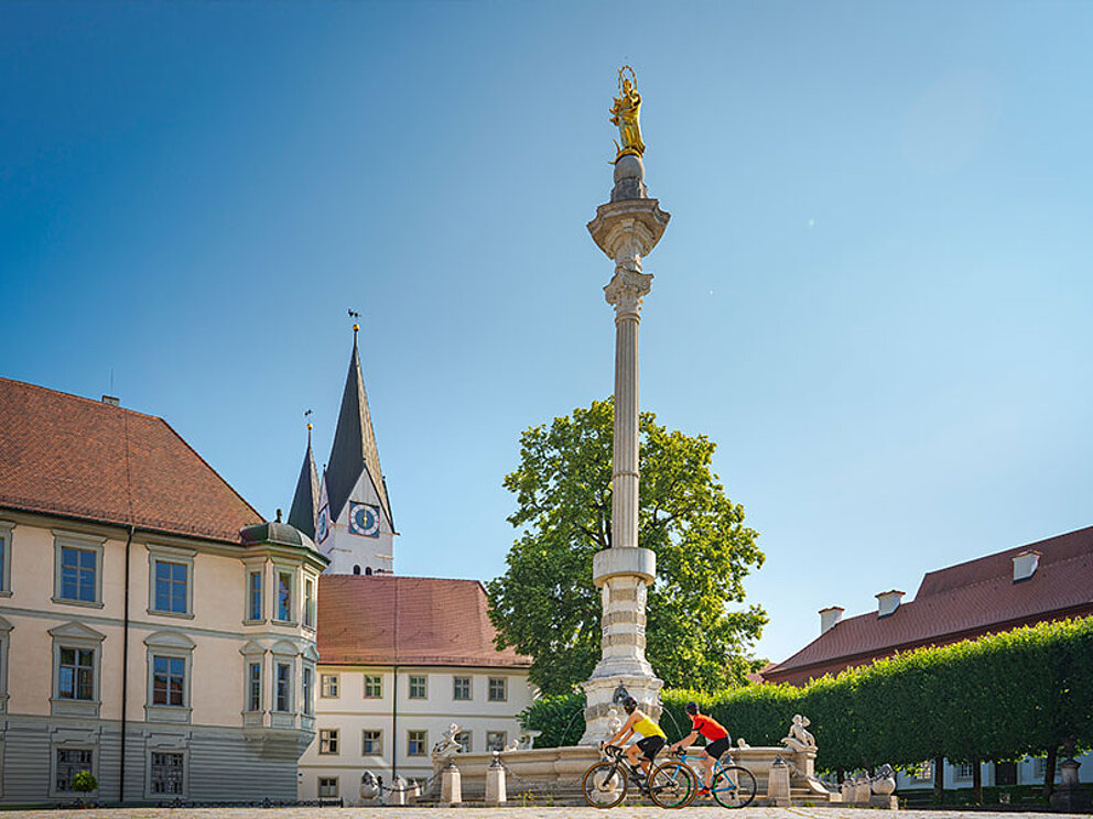 2 Radfahrer vor einem Brunnen, im Hintergrund ein Kirchturm