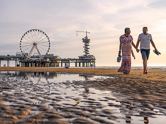 ein Paar geht Hand in Hand am Strand spazieren. Im Hintergrund ein Steg mit Riesenrad