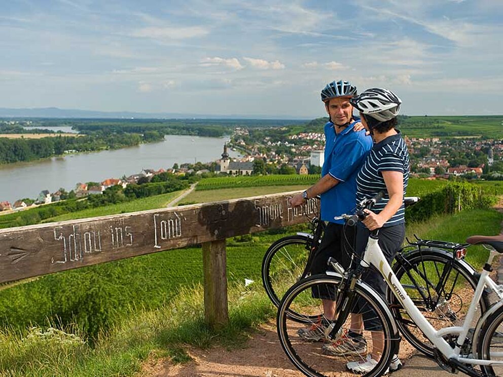 Eine Dame und ein Mann mit Radhelmen und Fahrrad blicken auf den Rhein. 