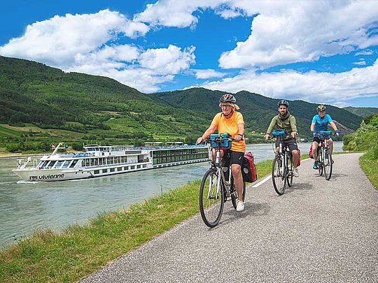 3 Radfahrer fahren am Radweg, im Hintergrund die MS Vivienne auf der Donau