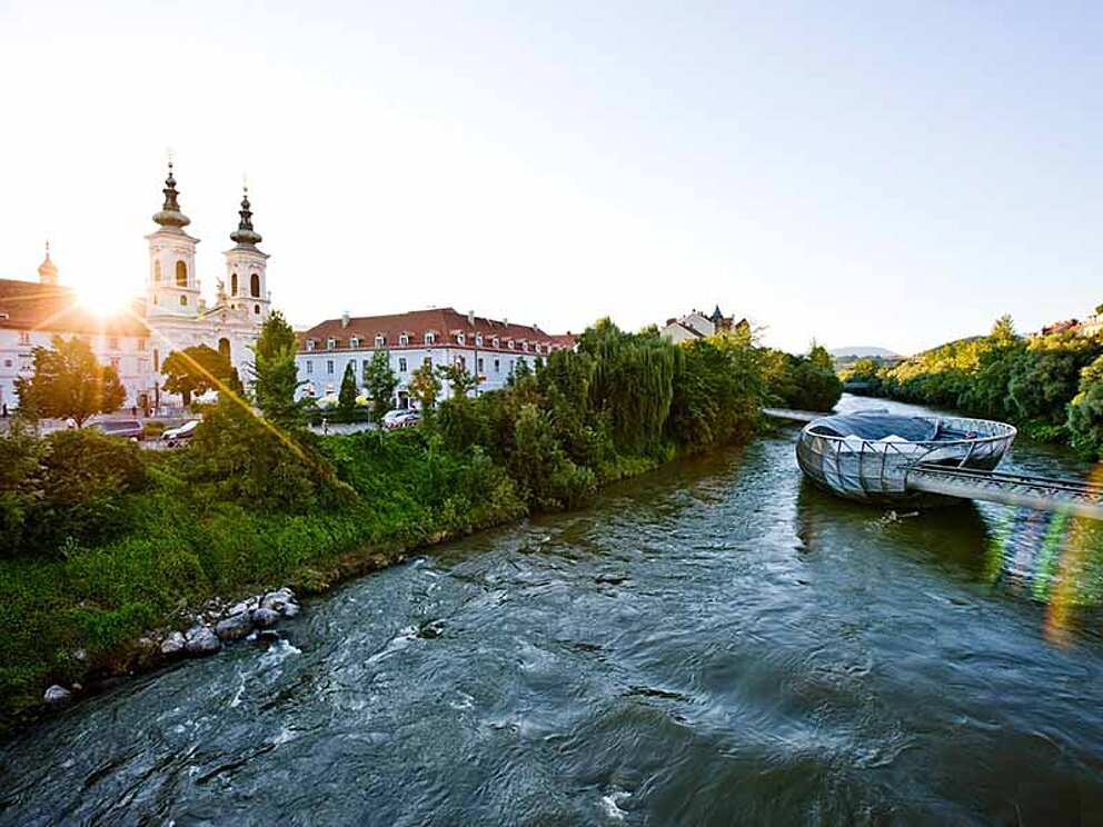 Murinsel mit untergehender Sonne hinter Mariahilfer Kirche in der Steiermark