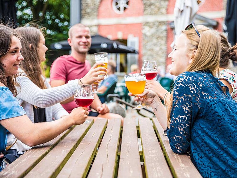 Eine Gruppe von Personen sitzt beim Tisch und prostet sich mit Bier zu.