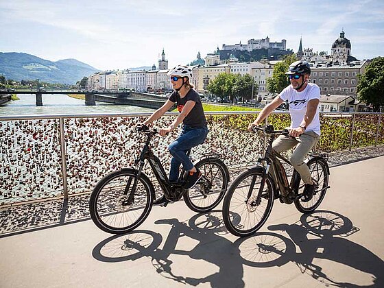 Mann und Frau auf E-Bikes fahren über die Salzachbrücke in Salzburg, dahinter die Festung