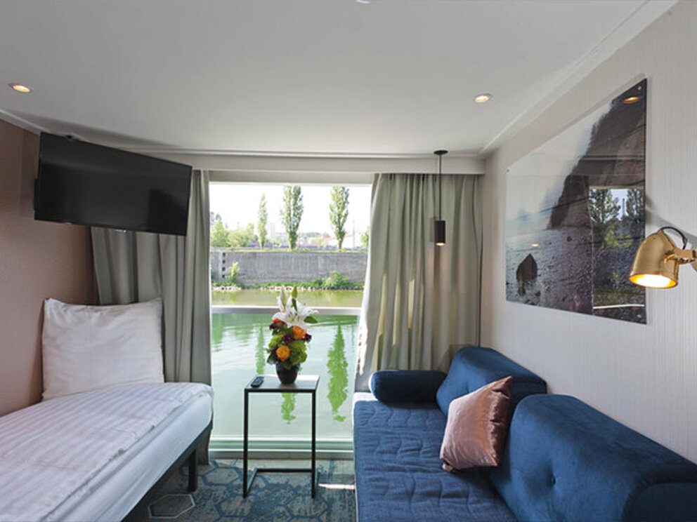 Kabine mit einem Einzelbett und einer gemütlichen Couch, bodentiefe Fenster mit Blick auf den Fluss.