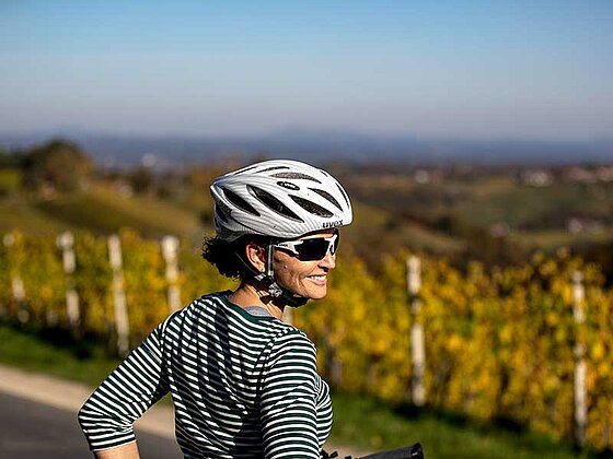 Eine Dame mit Fahrradhelm dreht sich zur Kamera. Im Hintergrund Weingärten.