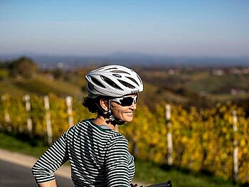Eine Dame mit Fahrradhelm dreht sich zur Kamera. Im Hintergrund Weingärten.