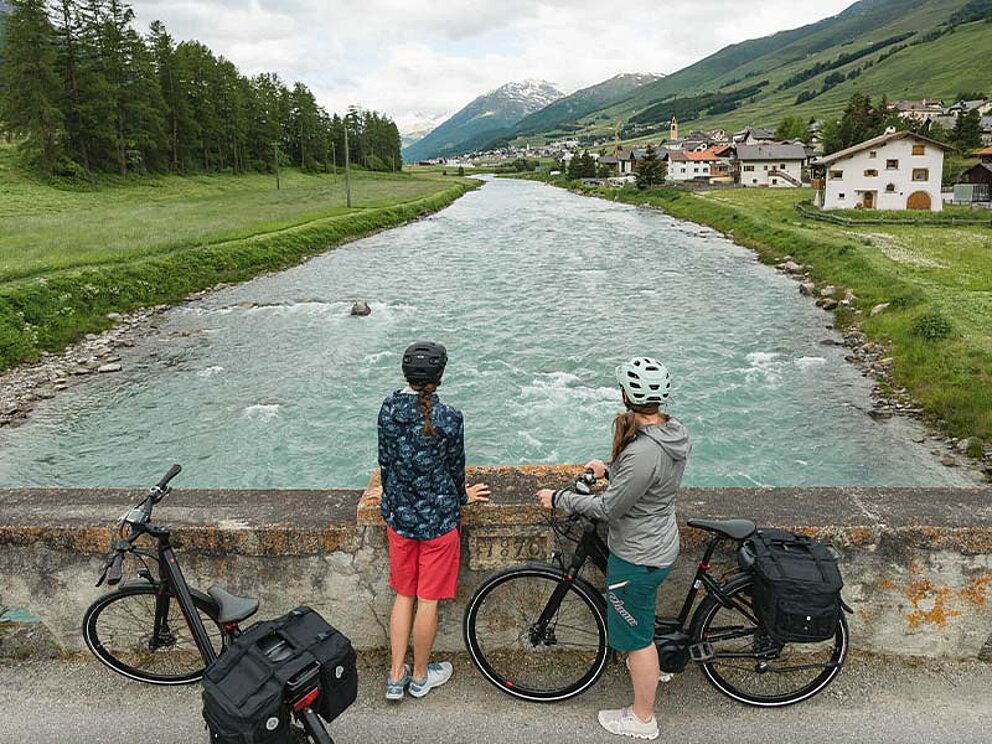 2 Radfahrer stehen auf einer Brücke und schauen in die Ferne über den darunterliegenden Fluss und die Berge