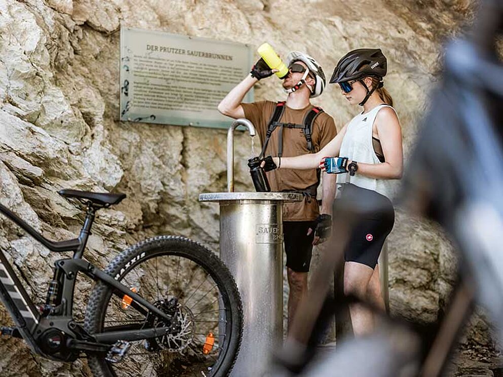 2 Mountainbiker füllen Ihre Trinkflaschen am Prutzer Sauerbrunnen mit mineralhältigem Trinkwasser