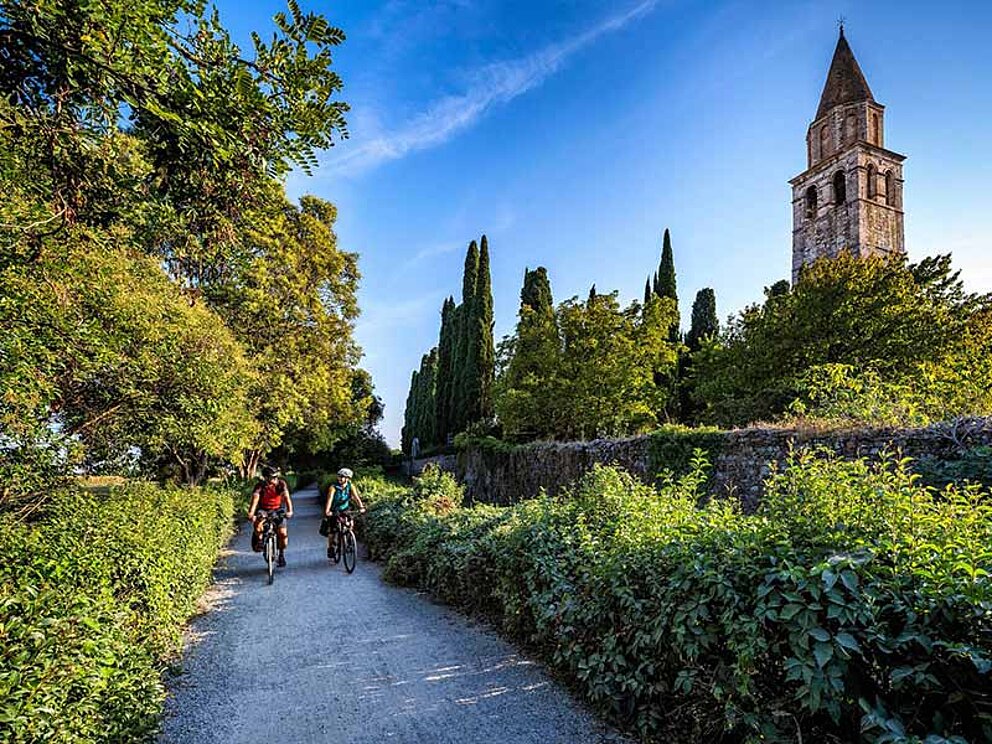 2 Radfahrer am Schotterweg, im Hintergrund eine Kirche