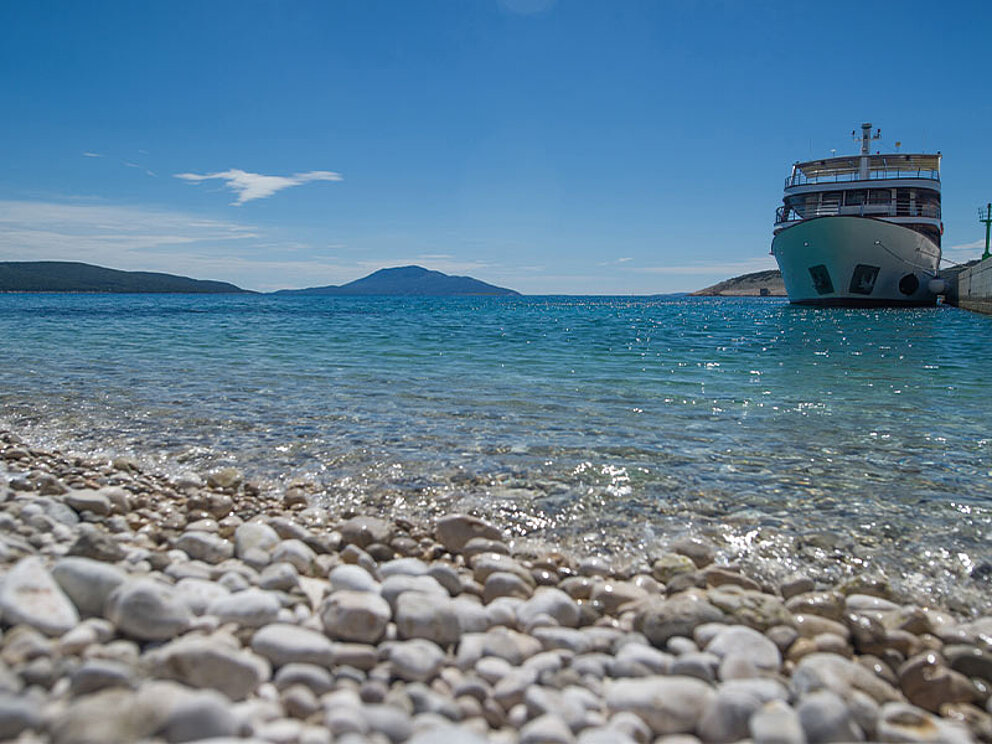 Ein Schiff liegt vor der Küste, im Vordergrund Kiesstrand und türkisblaues Meer
