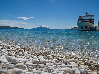 Ein Schiff liegt vor der Küste, im Vordergrund Kiesstrand und türkisblaues Meer