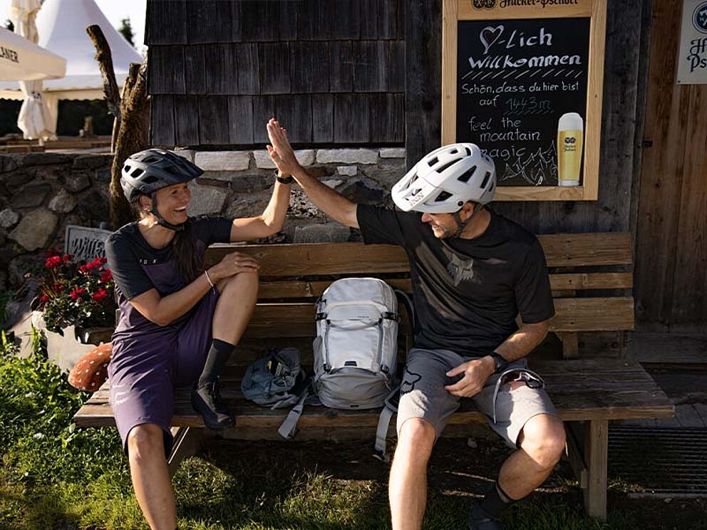 2 Biker auf einer Bank vor einer Almhütte geben high 5