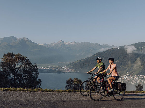 2 Radfahrer am asphaltierten Radweg, im Hintergrund der Zeller See und Berge