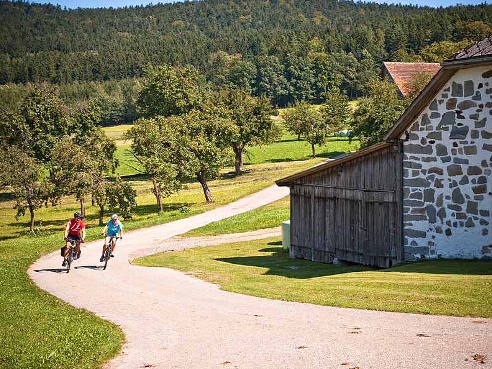 2 Radfahrer unterwegs am asphaltierten Radweg, vorbei an einem alten Bauernhof