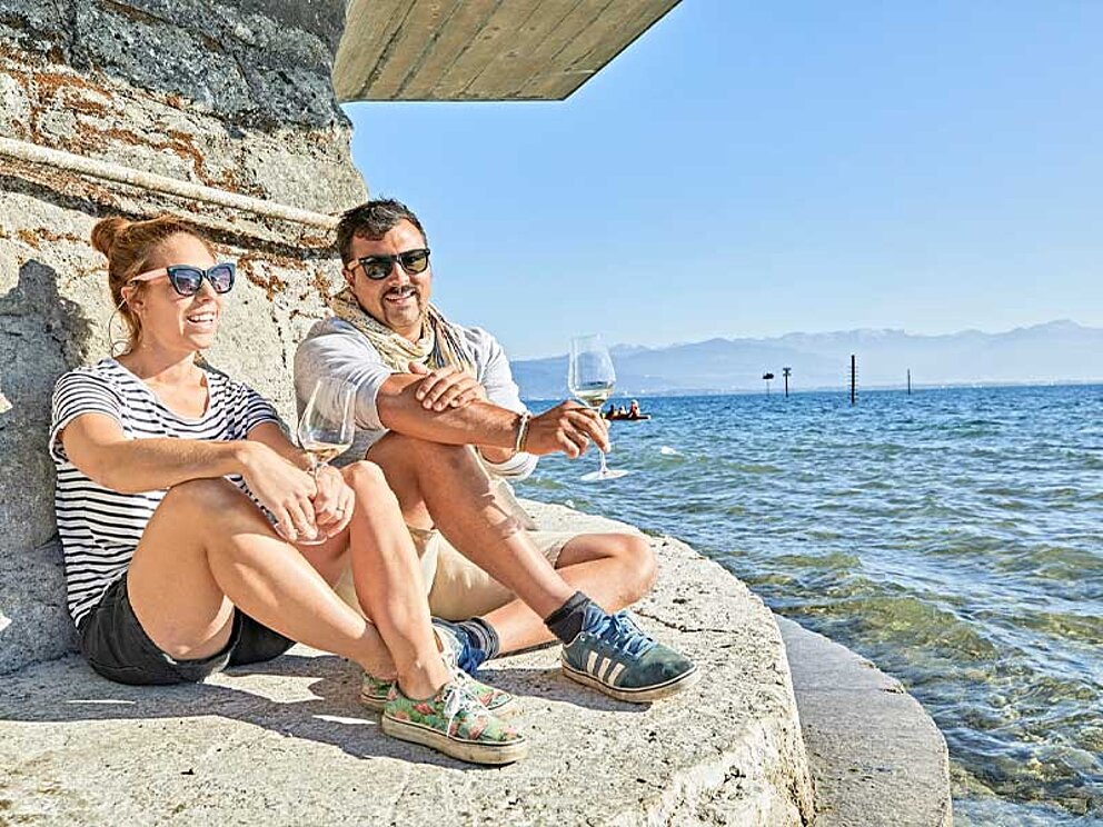 2 Personen sitzen auf einem Stein am Bodenseeufer mit einem Glas Weißwein in der Hand